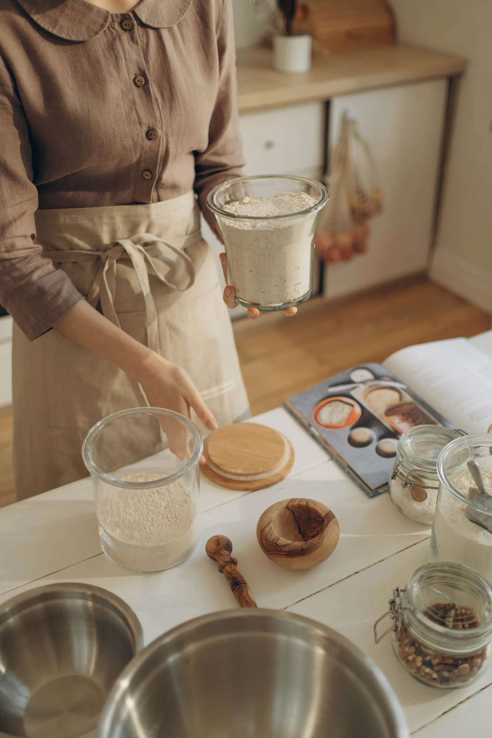 Ein Bäcker in Schürze hält ein Glas mit Mehl über einem Tisch, auf dem weitere Mehlgläser, ein Mörser mit Stößel und ein aufgeschlagenes Sauerteig-Rezeptbuch stehen