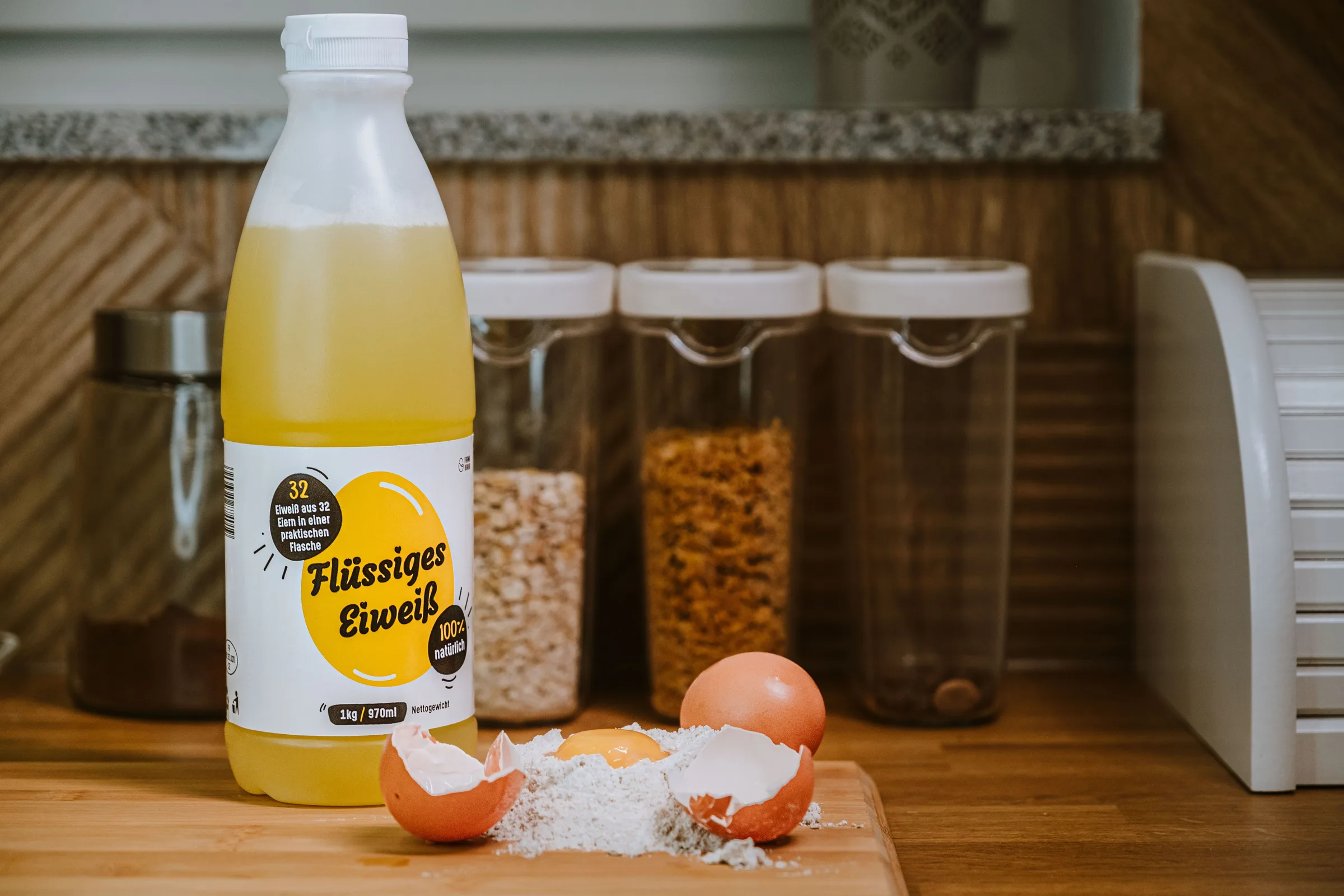 Flasche mit flüssigem Eiprodukt neben aufgeschlagenen Eiern und Mehl auf einer Küchenarbeitsplatte